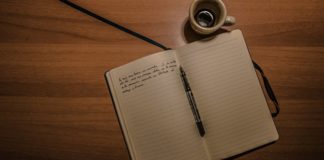 Imagem de divulgação. Tem um caderno aberto em branco com uma caneta e um tinteiro sobre uma mesa