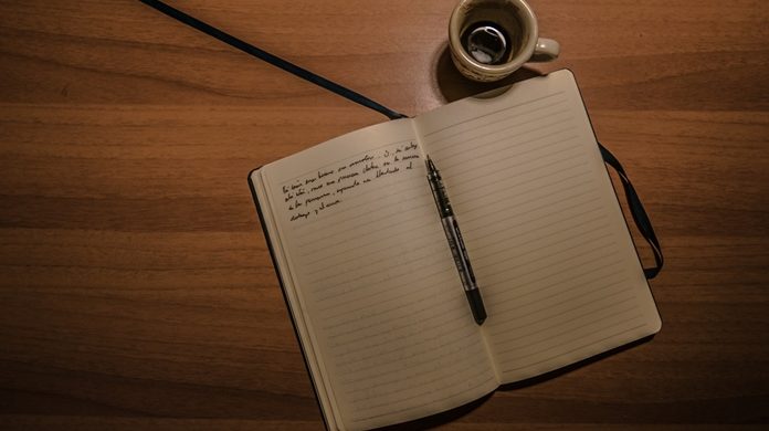 Imagem de divulgação. Tem um caderno aberto em branco com uma caneta e um tinteiro sobre uma mesa