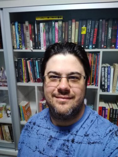 Rodrigo Basso, de camiseta azul, com óculos, no fundo um estante com livros
