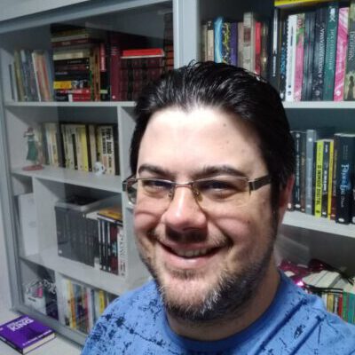 Selfie de Rodrigo Basso, sorrindo, usando óculos e camiseta azul. Um fundo com uma estante branca com vários livros