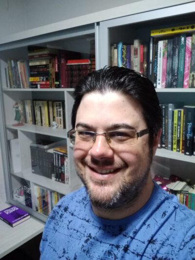 Selfie de Rodrigo Basso, sorrindo, usando óculos e camiseta azul. Um fundo com uma estante branca com vários livros