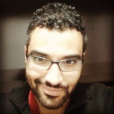 Elias Flamel, usando roupa escura, óculos e sorrindo