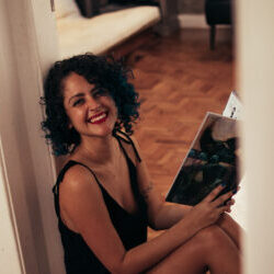 Ligia, sentada no chão, encostada na parede. Sorridente e segurando um livro