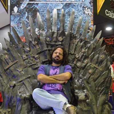 Paulo Vinicius sentado no trono de Game of Thrones, de braços cruzados, com camiseta roxa e calça jeans, com perna cruzada