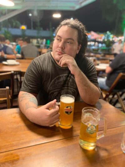 Samuel Muca sentado em uma mesa de bar, com mão apoiada na mesa segurando uma caneca com cerveja