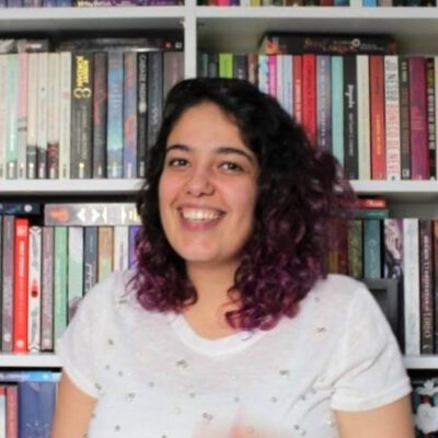 Taina, sorridente, com um cabelo preto com pontas roxas, usando uma blusa branca. No fundo uma estante cheia de livros