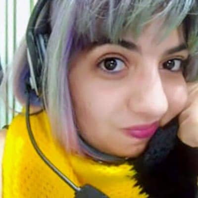Selfie de Aline Bergamo. Mulher branca, cabelo loiro com mechas roxas e verdes, com a mão apoiada no queixo. Está usando um headset preto e vestindo uma regata preta e um cachecol amarelo.