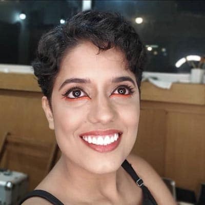 Selfie de Carol Lima. Uma moça parda e cabelos curtos e cacheados sorri olhando para cima. Nos olhos um delineador laranja e na boca batom avermelhado.