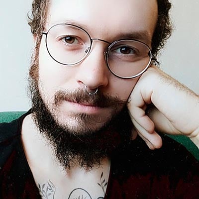 Selfie de Franco Wolff. Homem branco, com cabelo e barba castanha, com a mão apoiada no rosto. Está usando óculos e vestindo uma camiseta preta.