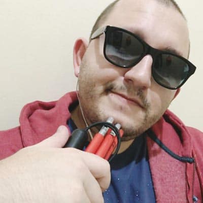 Selfie de Sidney Andrade. Homem branco, cabelos raspados, com barba. Está usando óculos escuros e vestindo um moletom vermelho, enquanto segura uma bengala dobrável vermelha.