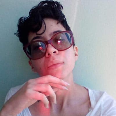 Selfie de Yohanne Zamboti. Mulher branca, cabelo preto e curto, com a mão apoiada no queixo. Está usando óculos escuros e vestindo uma camiseta branca.