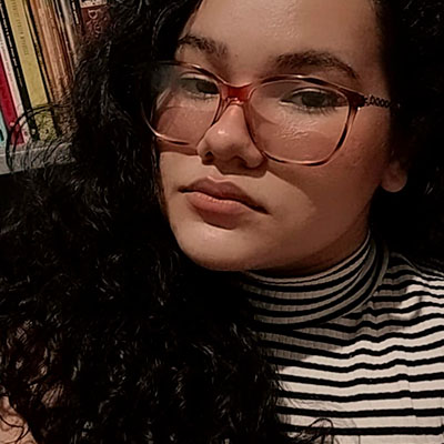 Mulher de cabelos pretos ondulados, usa óculos com armação vinho e blusa listrada está em frente a uma estante de livros