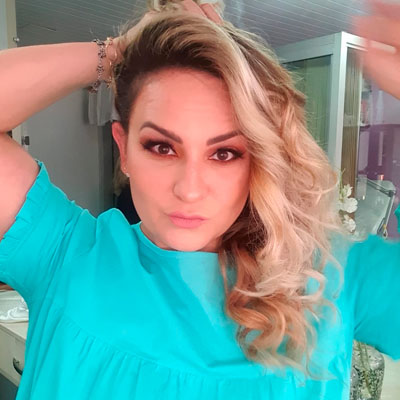 Mulher branca com cabelo longo, loiro descolorido e raiz aparente. A mão direita sobre a cabeça, mostrando uma pulseira, e usando uma blusa branca.