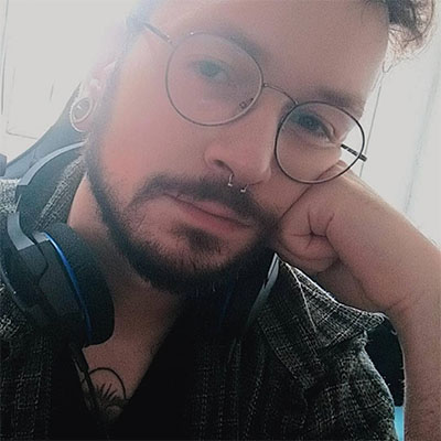 Homem branco com barba curta, óculos de armação redonda e cinza, piercing cinza no septo, cabelo curto e a bochecha esquerda apoiada na mão esquerda, com um headset preto no pescoço e usando uma camiseta preta com uma camisa xadrez verde acinzentada.