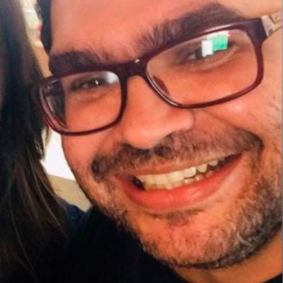 Homem branco na casa dos 30 anos, usando óculos vermelho, com barba rala e sorrindo.