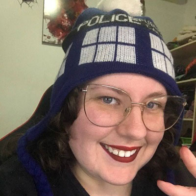 Mulher branca de batom vermelho sorrindo, vestindo camiseta preta, óculos de armação dourada e um gorro azul de Doctor Who, fazendo um V com os dedos da mão.