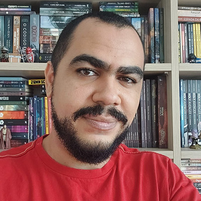 Selfie de Ace Barros. De cabelo e barba escuros e bem curtos, usando uma camiseta vermelha. No fundo uma estante repleta de livros.