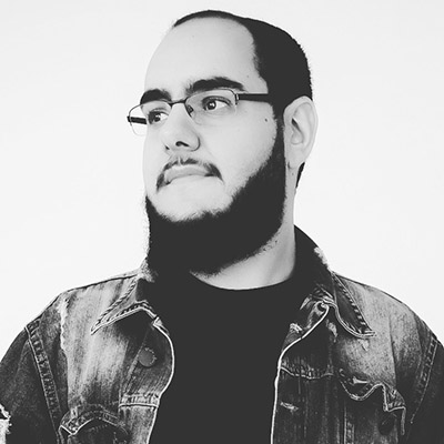 Imagem, em preto e branco, de rosto de Leonardo. Ele usa barba escura, óculos, uma camiseta preta e jaqueta jeans. Ele está virado para frente olhando o lado.